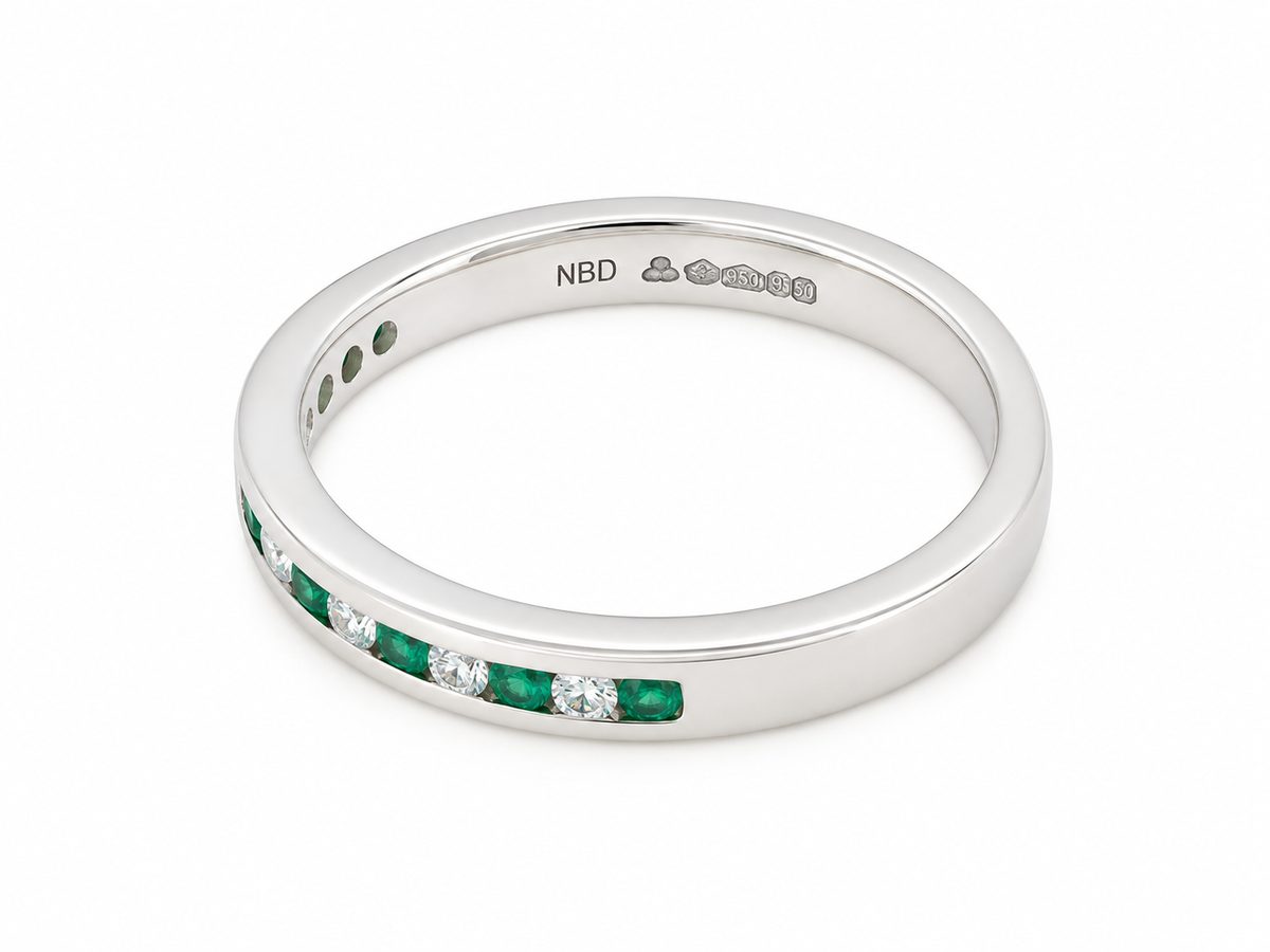 An eternity-style band ring featuring a channel setting of green emerald-cut gemstones and clear round stones, shown on a clean white background. The inside of the band is stamped with 'NBD'.